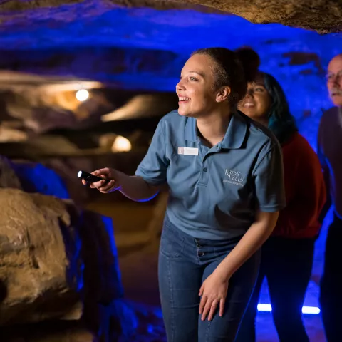 Tour guide in blue shirt and flashlight leads small group through cavern